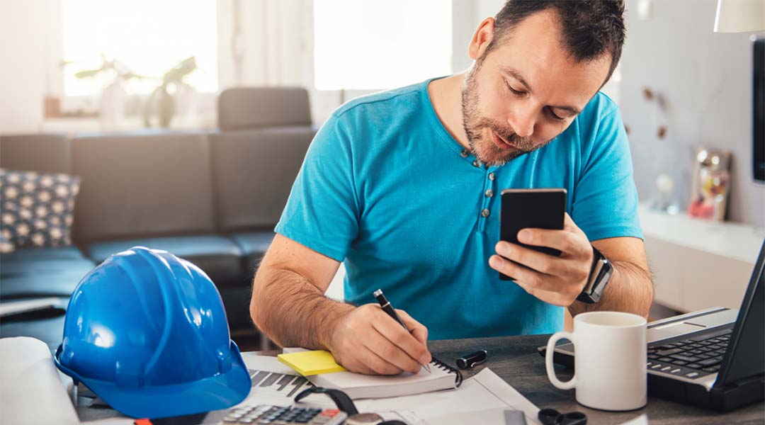 Man in blue shirt writing notes and holding smart phone at home office